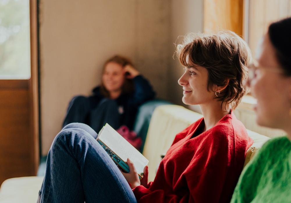 Drie jongeren zitten ontspannen in een woonkamer. De persoon op de voorgrond draagt een groene trui, de persoon in het midden draagt een rode trui en houdt een boek vast terwijl ze glimlacht, en op de achtergrond zit iemand nonchalant tegen de muur geleund. Het daglicht valt door een raam naar binnen.