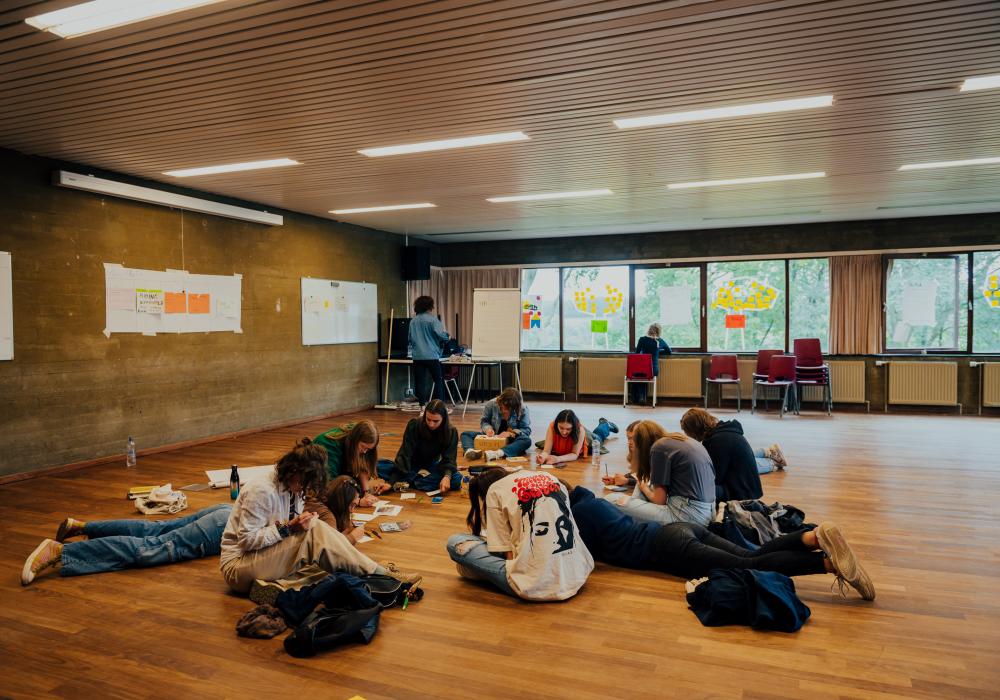 Een groep jongeren zit en ligt op de vloer van een grote zaal met houten vloer, terwijl ze creatief bezig zijn met papier en stiften. Aan de muren hangen posters en whiteboards met aantekeningen. Op de achtergrond werken twee mensen aan bureaus bij een raam met kleurrijke post-its.