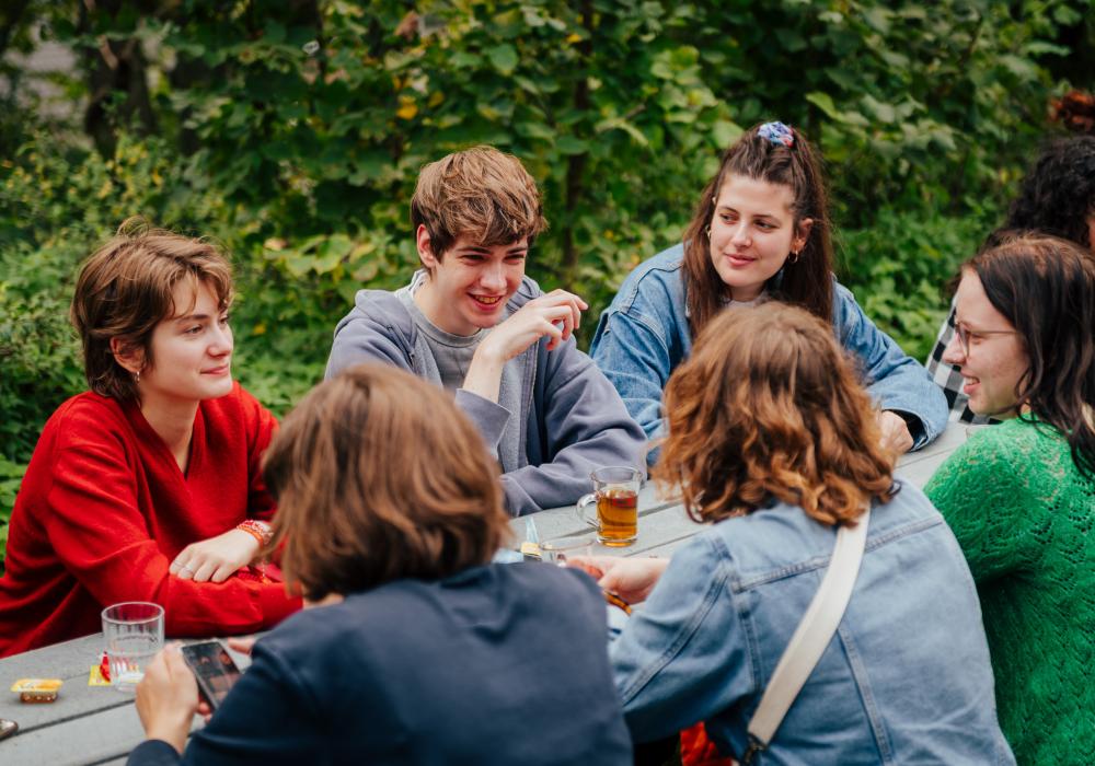 Een groep jongeren zit buiten aan een picknicktafel, omringd door groen. Ze praten, lachen en drinken thee, terwijl sommigen op hun telefoon kijken. De sfeer is ontspannen en gezellig.