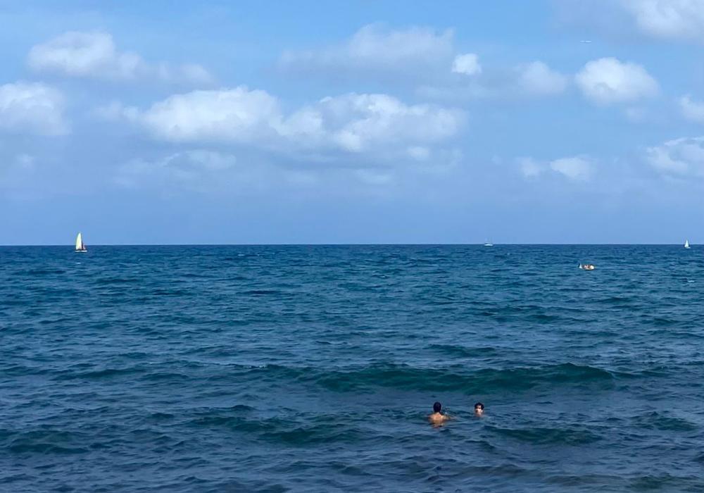 Foto van tijdens een DiscoverEU-reis: de zee, blauwe hemel met een paar wolkjes, twee jongeren zwemmen in de zee