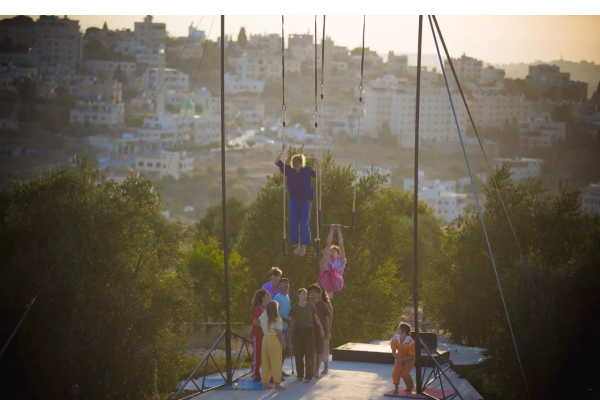 Een groep jonge circusartiesten staat op een verhoogd platform buiten met een stad op de achtergrond. Twee mensen hangen aan trapezes boven het platform, terwijl anderen toekijken. De zon schijnt helder.