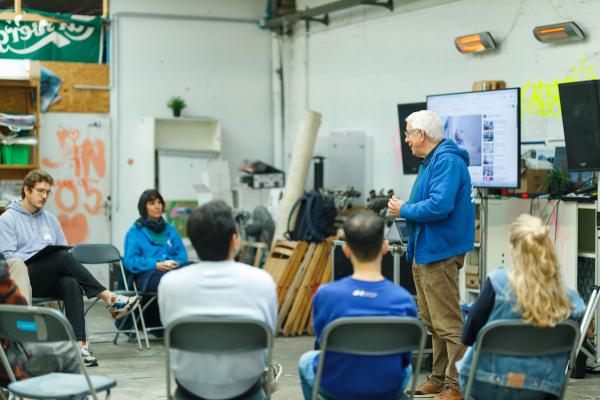 Ton geeft een presentatie aan een kleine groep mensen die in een halve cirkel zit, in een informele ruimte met industrieel karakter.