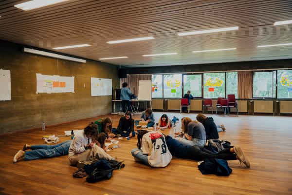 Een groep jongeren zit en ligt op de vloer van een grote zaal met houten vloer, terwijl ze creatief bezig zijn met papier en stiften. Aan de muren hangen posters en whiteboards met aantekeningen. Op de achtergrond werken twee mensen aan bureaus bij een raam met kleurrijke post-its.