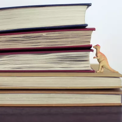 Afbeelding van een dino die in boeken kijkt voor de Jeugdboekenmaand met thema tijd Jeugdboekenmaand thema 2025 tijd