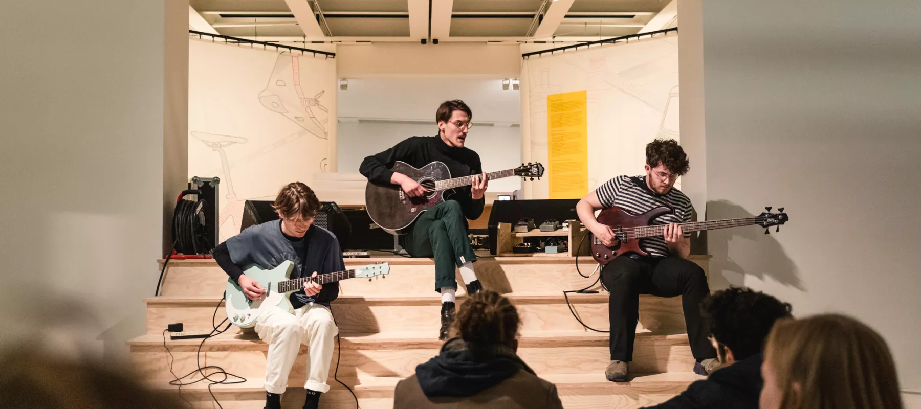 Drie mannen op een trap spelen elektrische gitaar omringd door 2 schilderijen