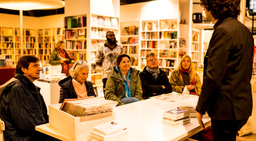 Een groepje mensen rond een tafel in een boekhandel, luisterend naar een auteur