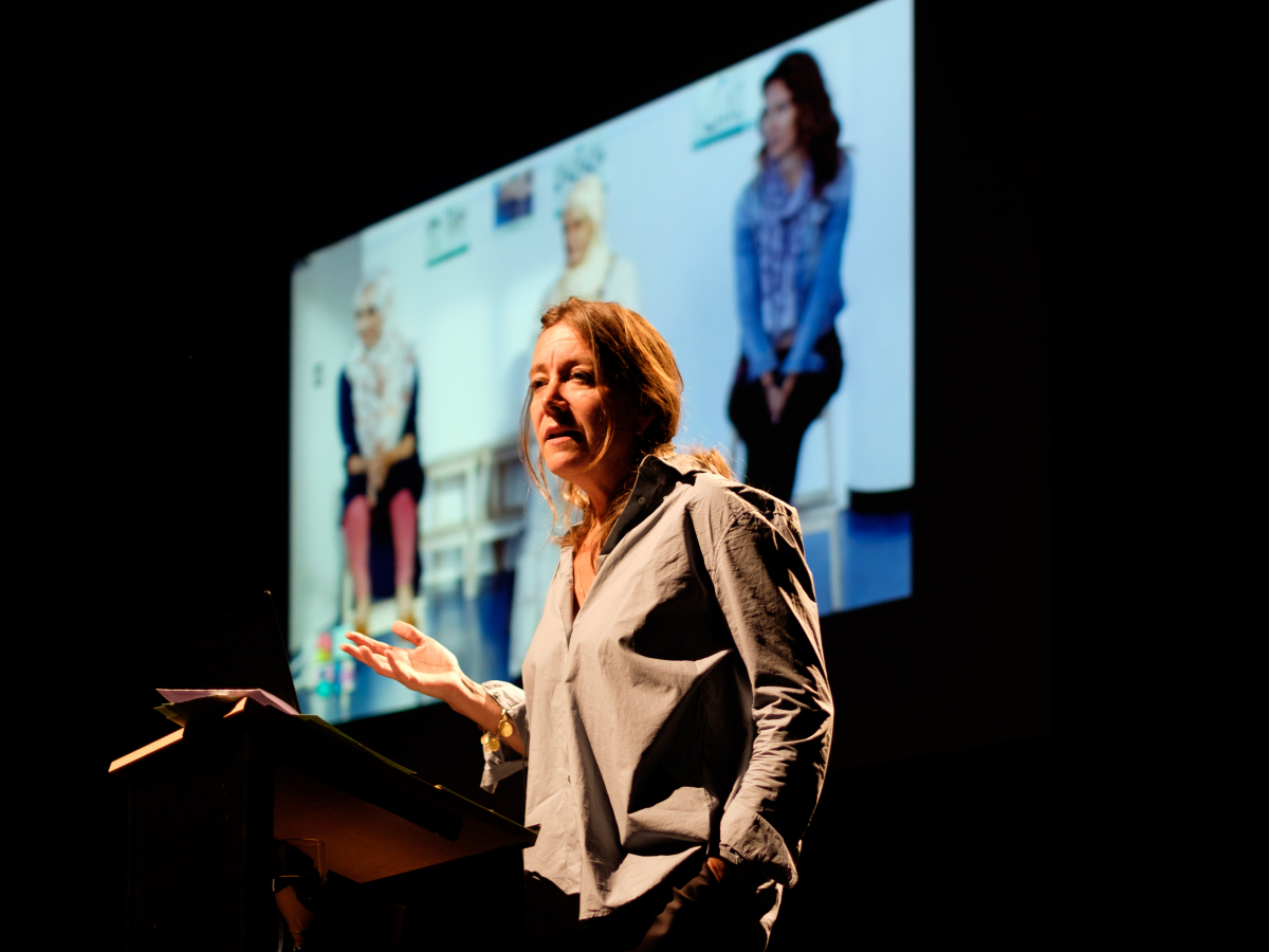 Een vrouw die uitleg geeft met een powerpointpresentatie op de achtergrond.