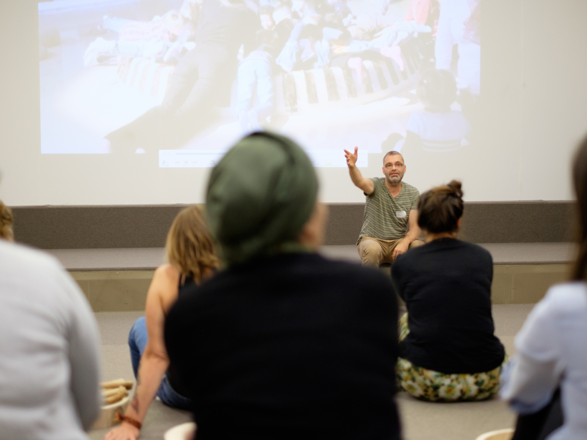 Een zittend persoon die aan een groep mensen een lezing geeft met een geprojecteerd beeld op de achtergrond.